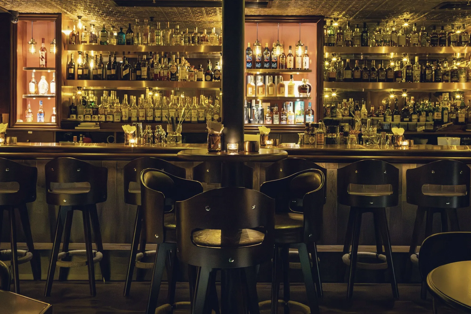 A dimly lit cocktail bar with backlit liquor shelves and a row of empty stools.