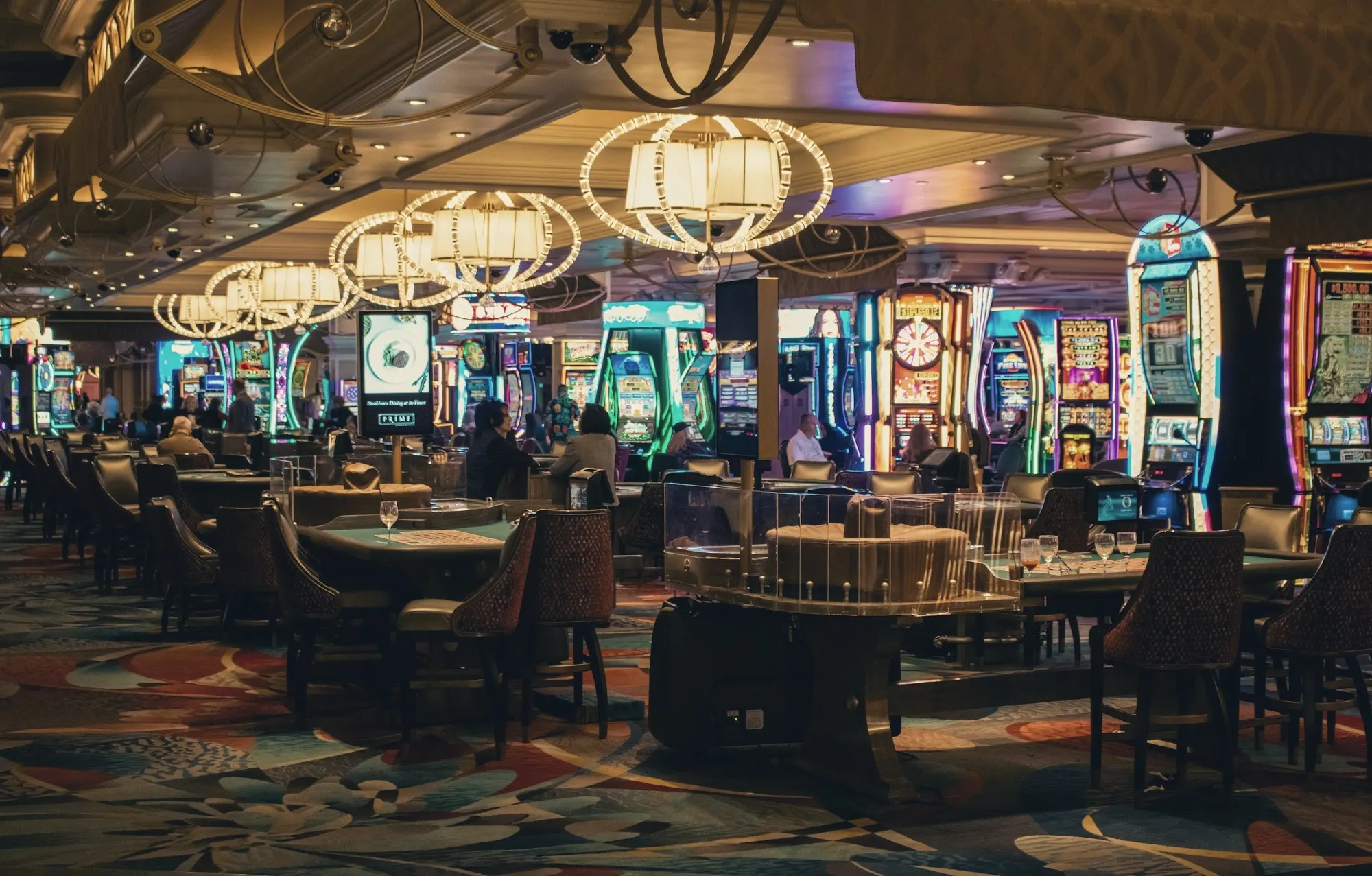 A casino floor with card tables, slot machines and ornate chandeliers under a low ceiling.