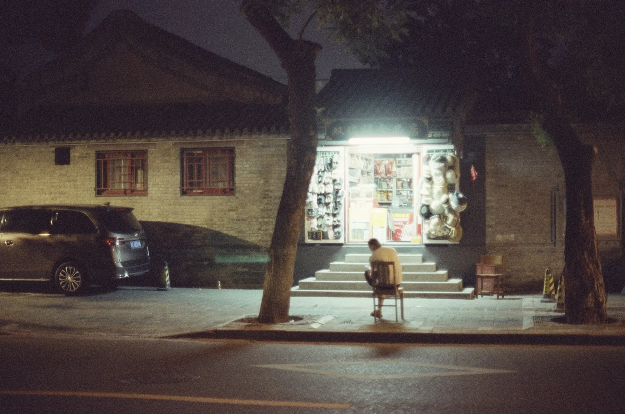 A brightly lit corner shop at night on a quiet street with a parked car.