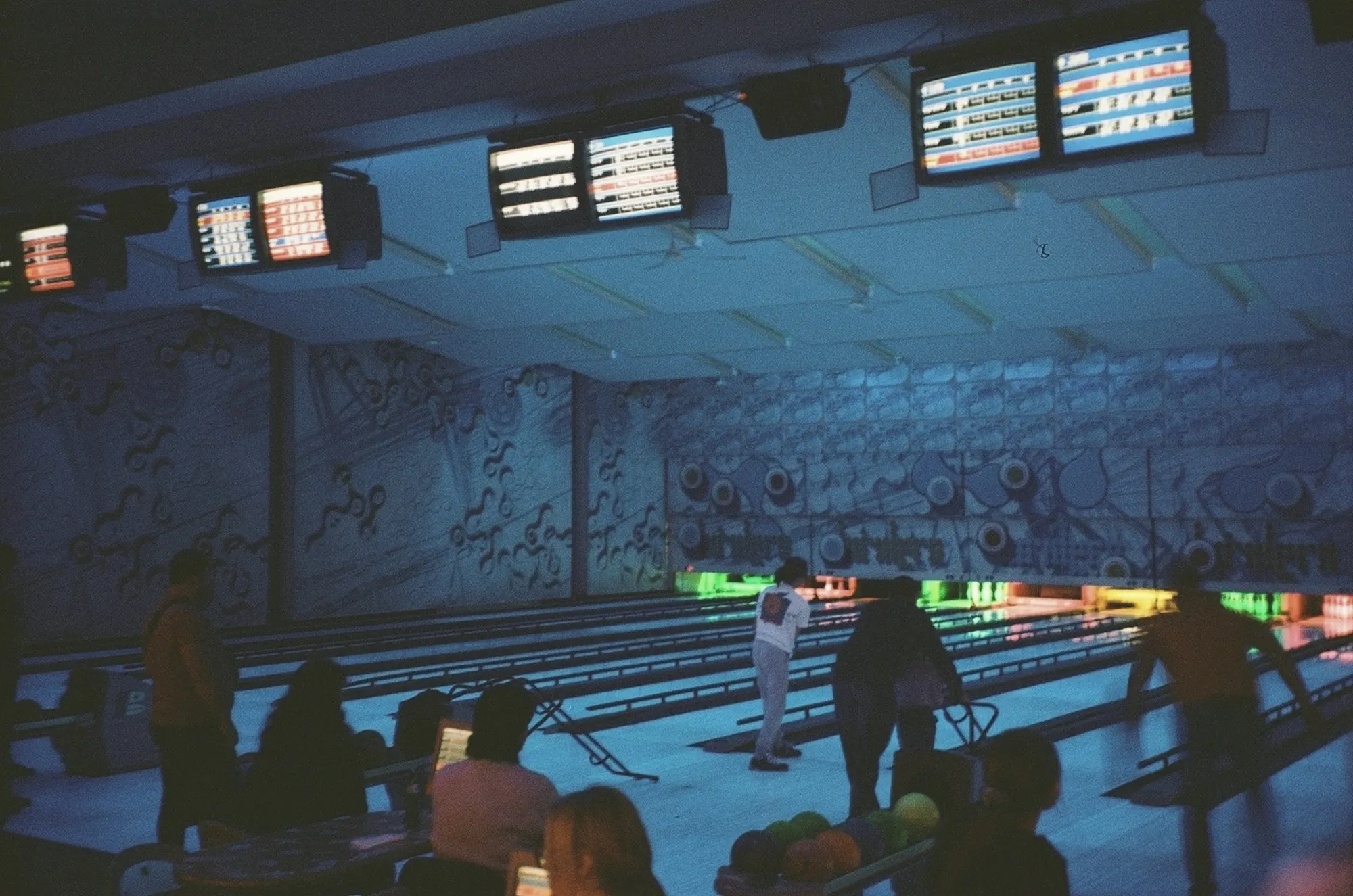 Bowlers on glowing lanes in a bowling alley under overhead score monitors.