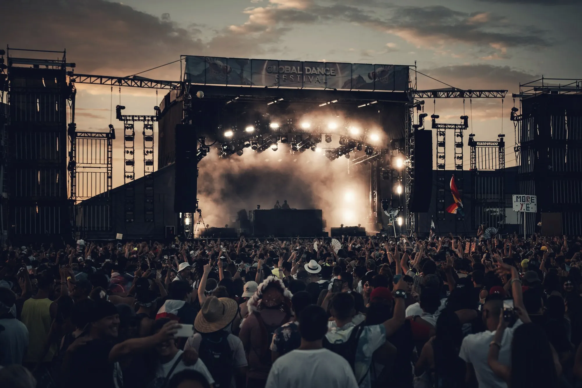 A large crowd facing a lit festival stage with smoke and lighting rigs at dusk.