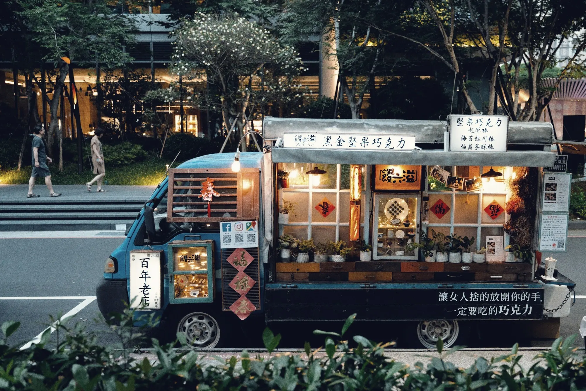 A decorated food truck with a lit serving window parked on a city street at dusk.