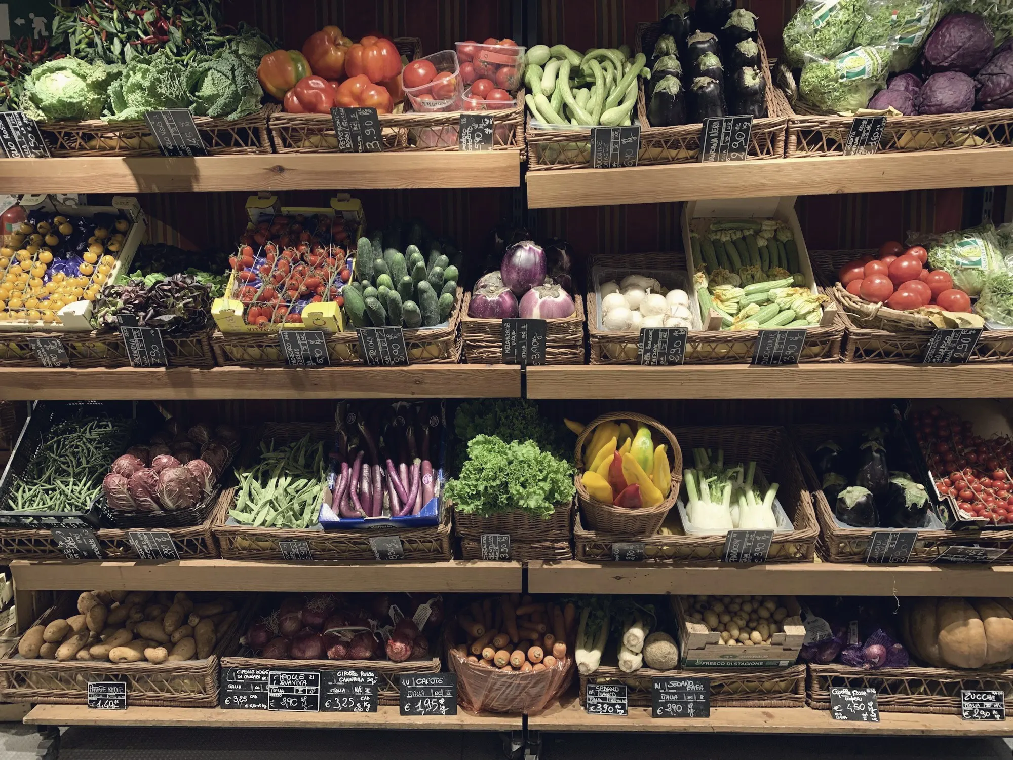 Wooden shelves stocked with baskets of fresh vegetables and handwritten price tags.