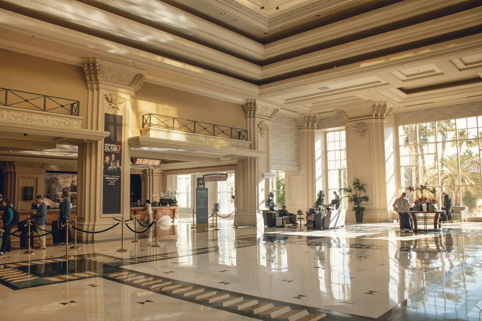 A sunlit hotel lobby with marble floors, tall columns and guests at the front desk.