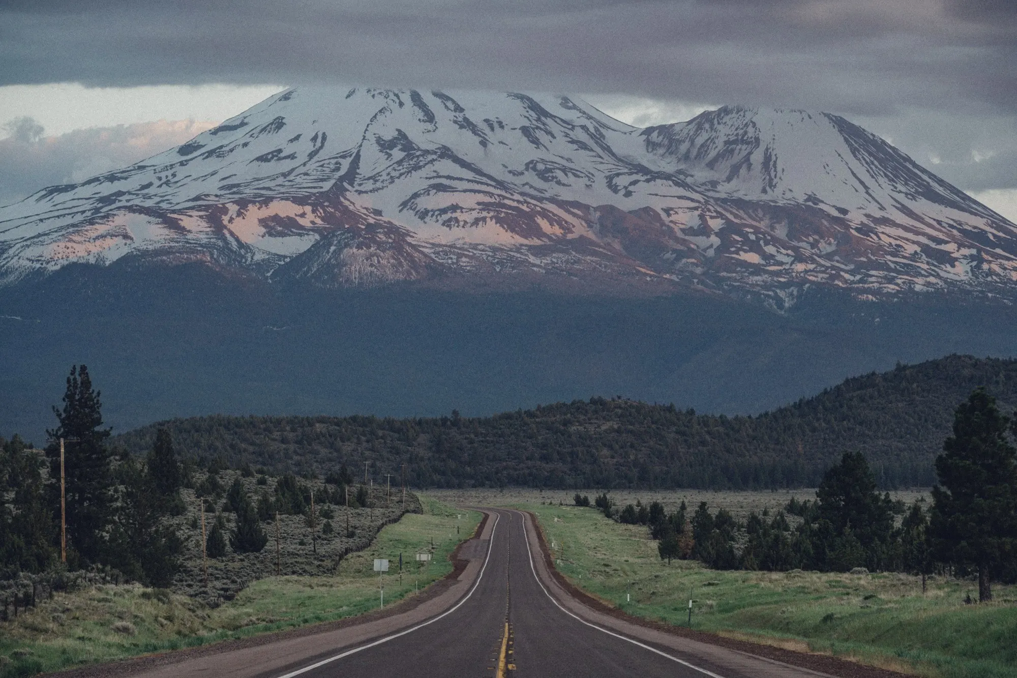 A straight two-lane road leading toward snow-covered Mount Shasta under cloudy skies.