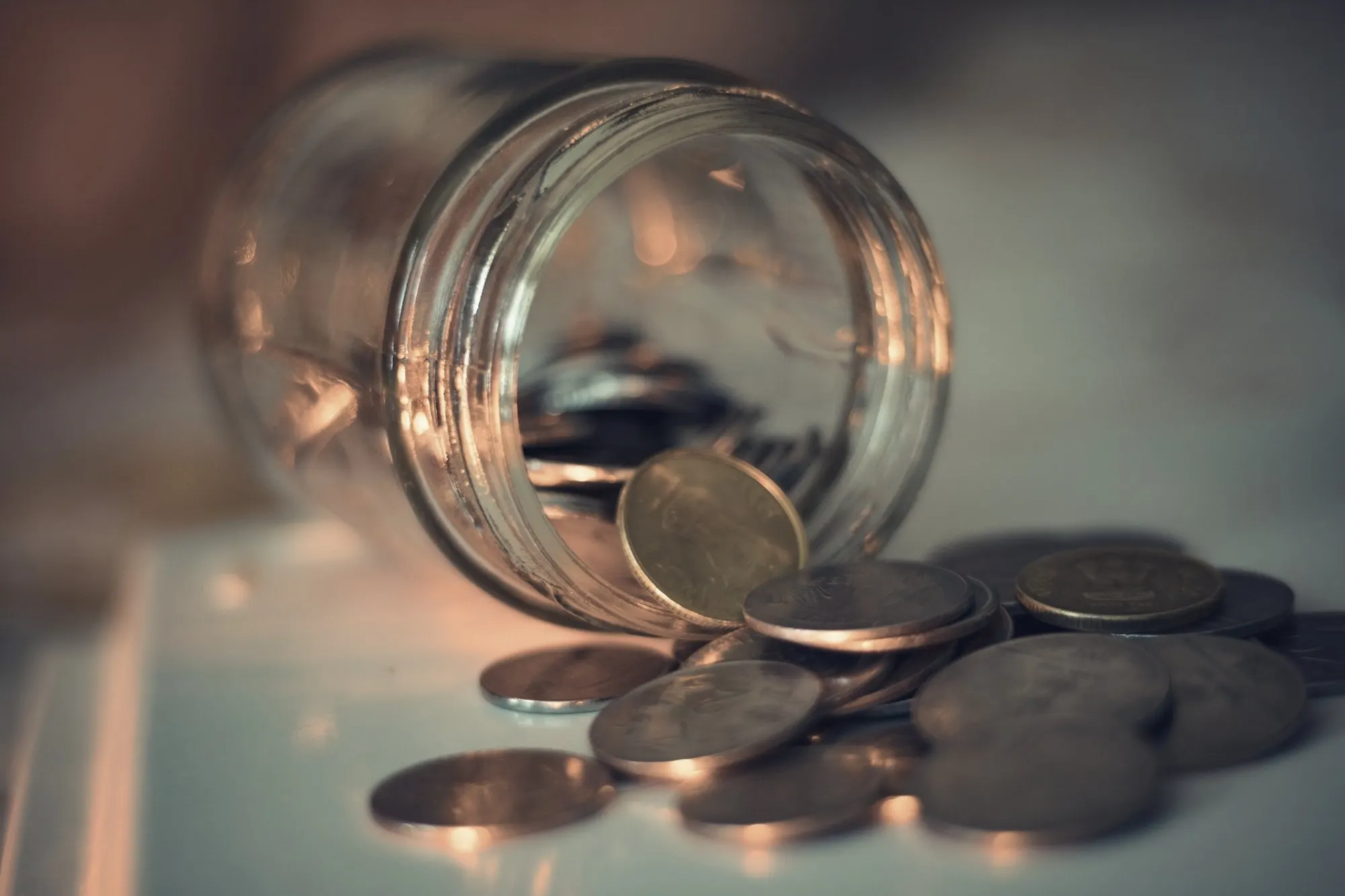 A tipped glass jar spills silver and copper coins across a table in warm light.
