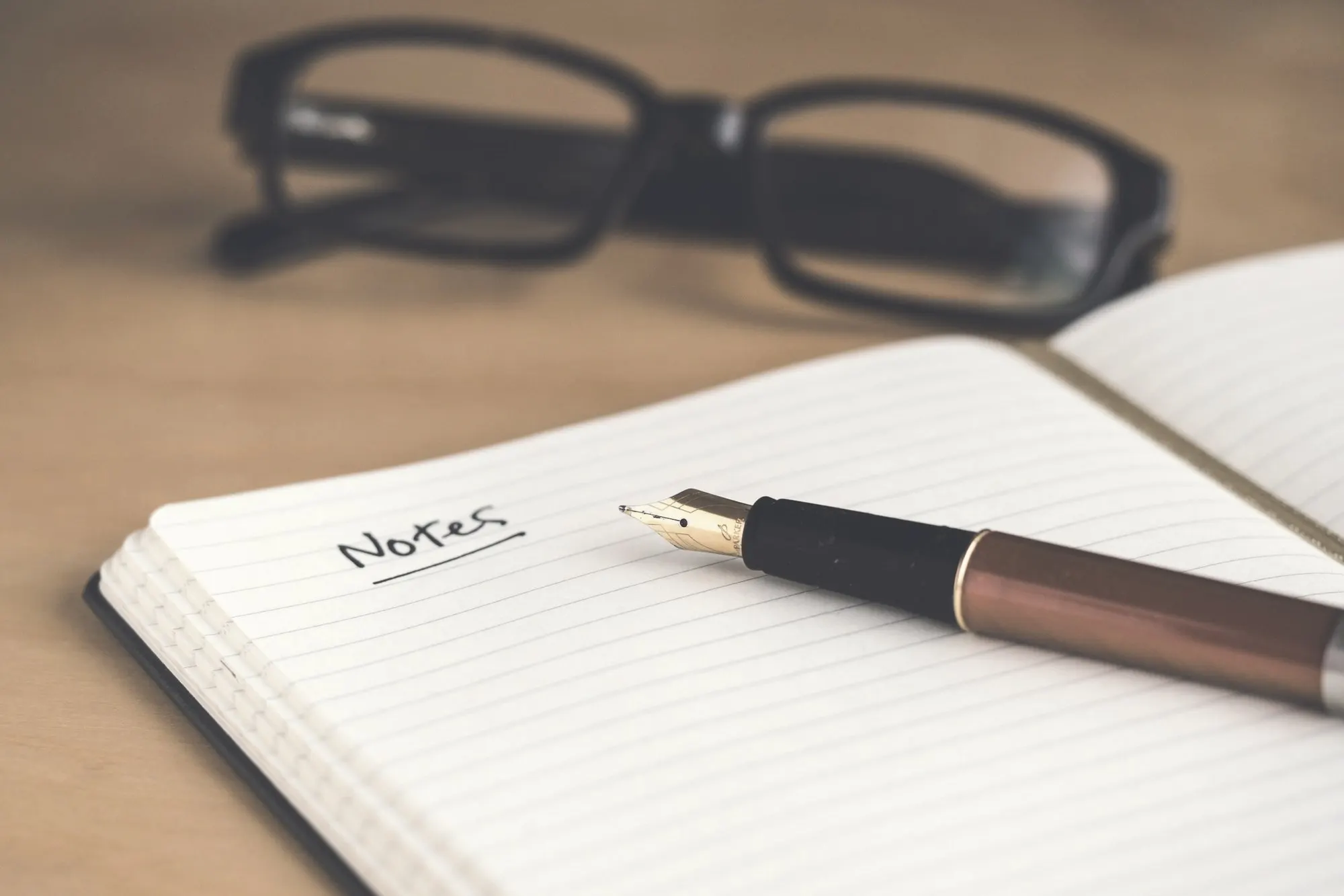 Fountain pen with gold trim lying on an open lined notebook beside black glasses.