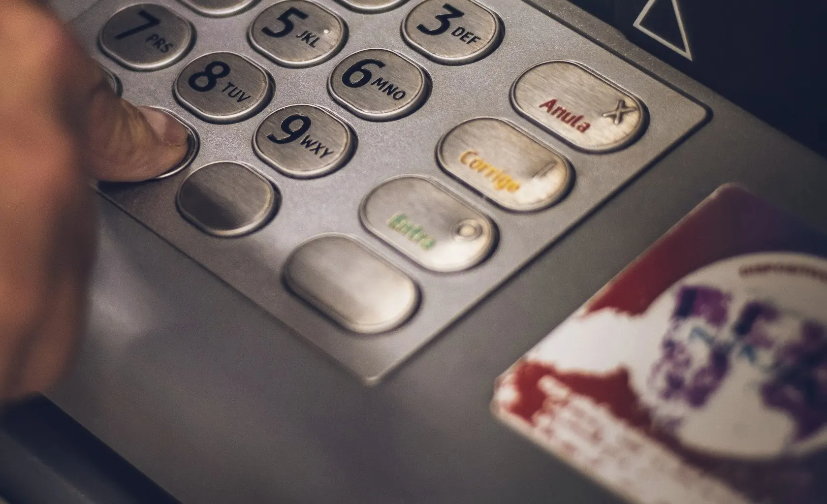 A finger presses the metal keypad buttons on a cash machine in low light.
