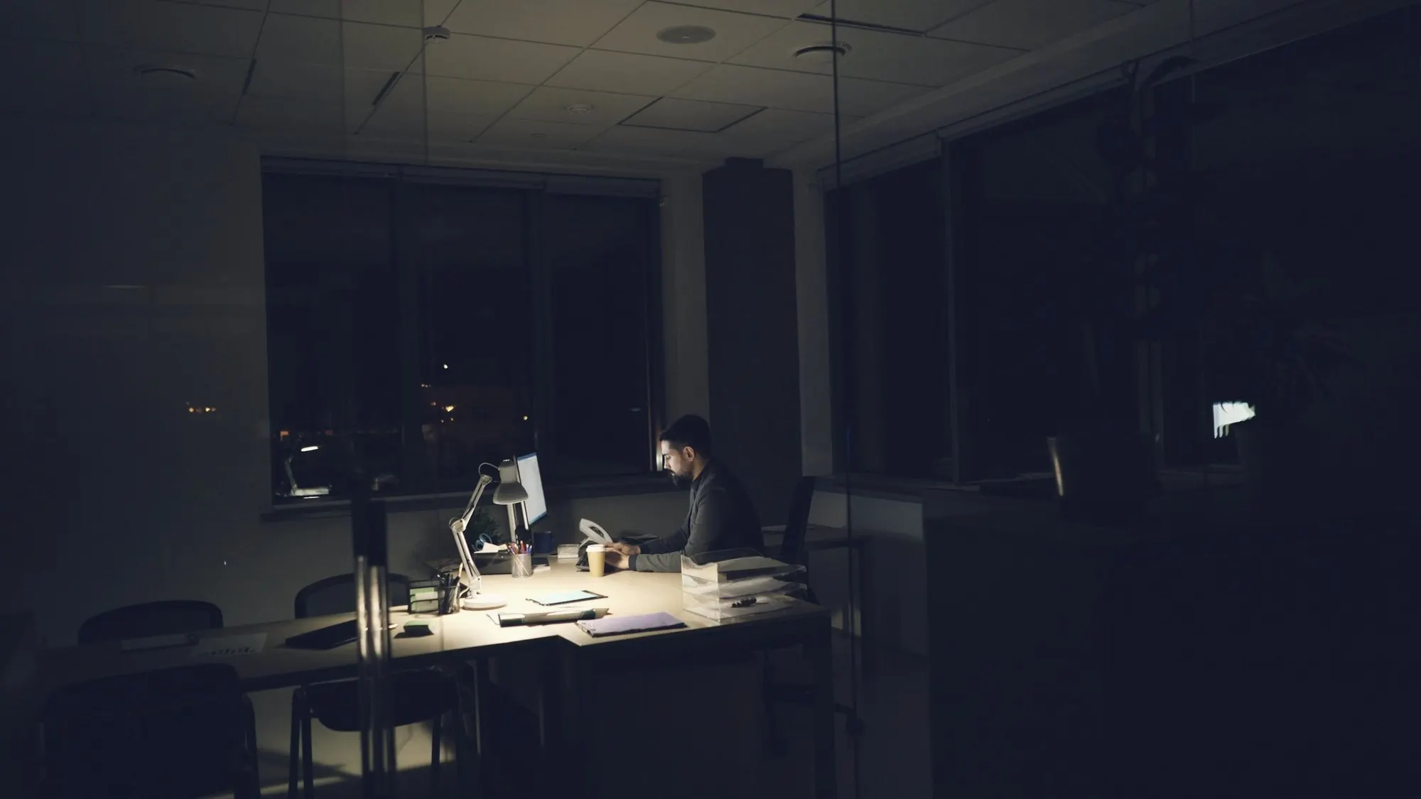 A single desk lamp lights a late-night office where one person works.