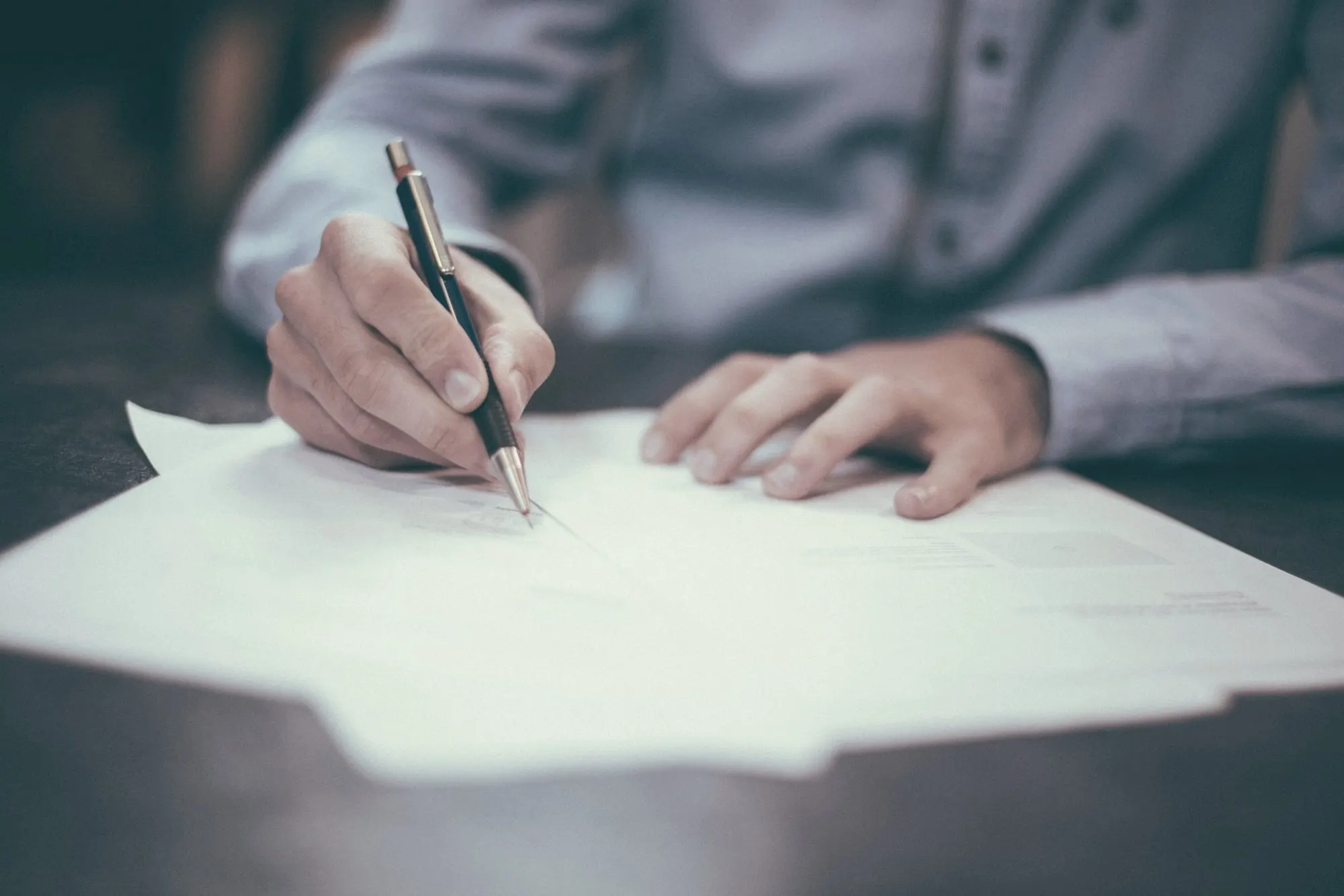 A person in a blue shirt signs a paper document with a pen at a desk.