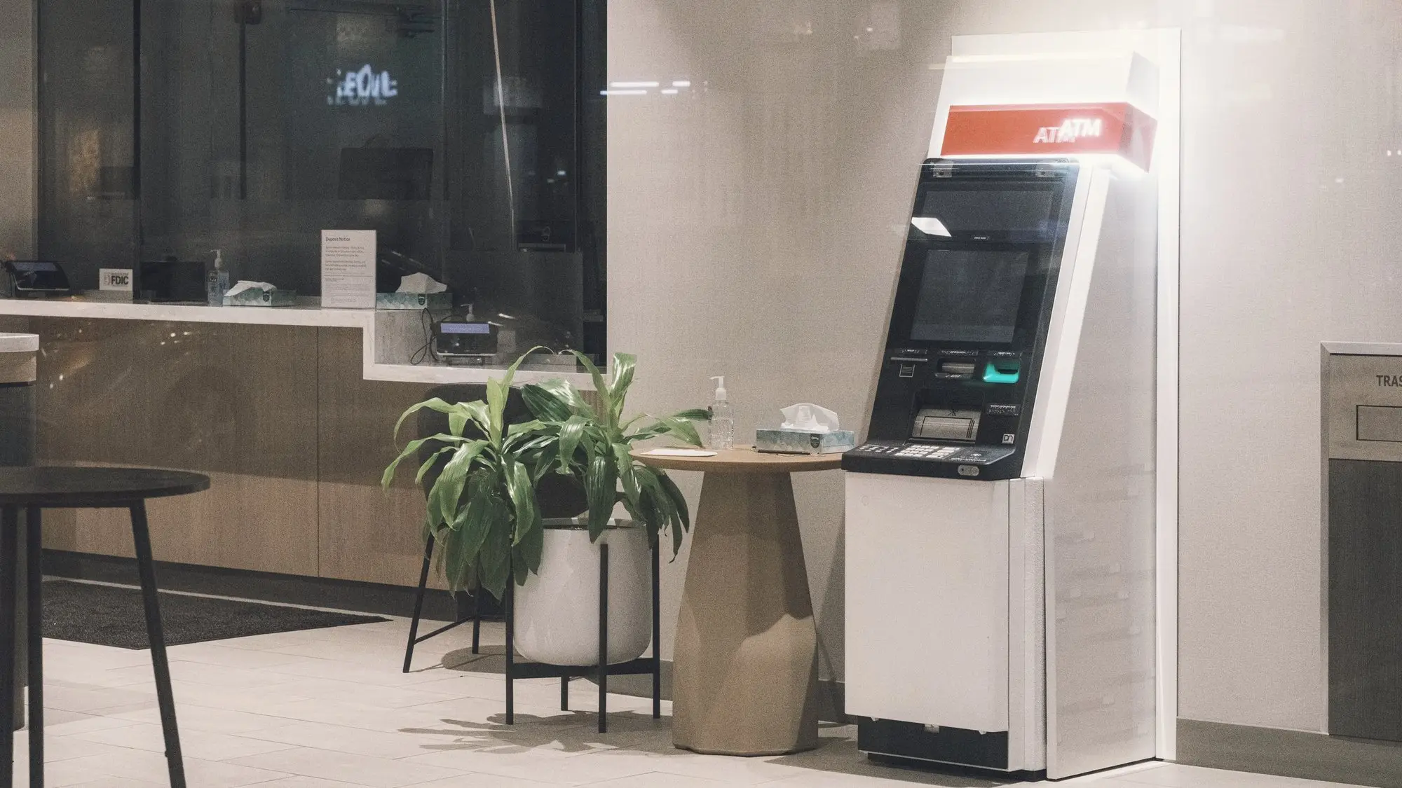 A freestanding ATM in a quiet, modern lobby, next to a potted plant and a small round table.