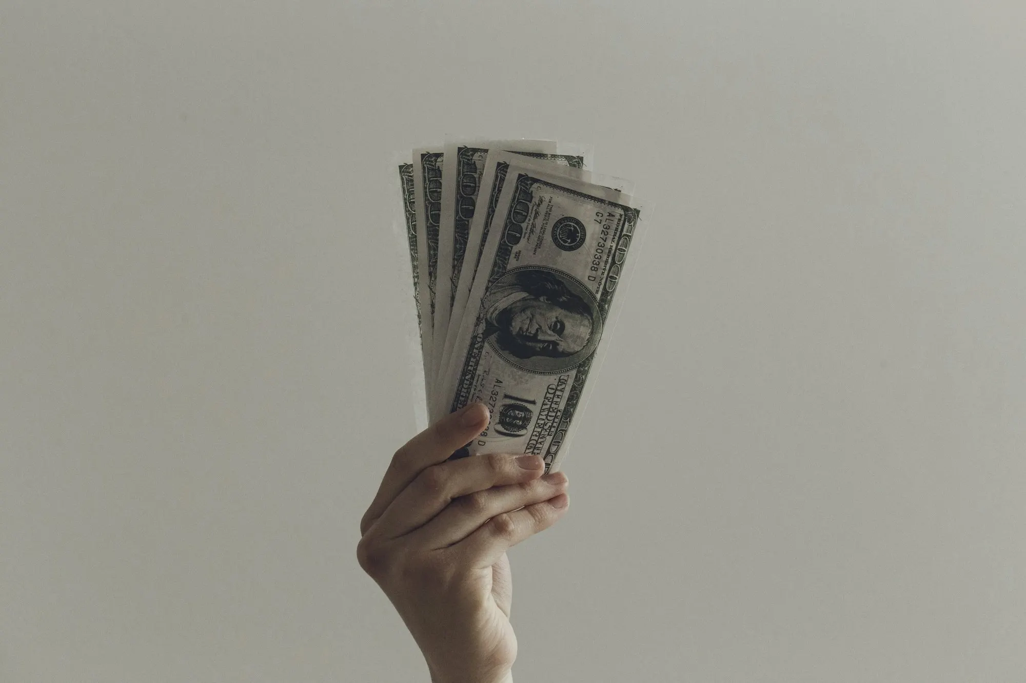 A hand holding a fan of dollar bills against a plain gray wall.